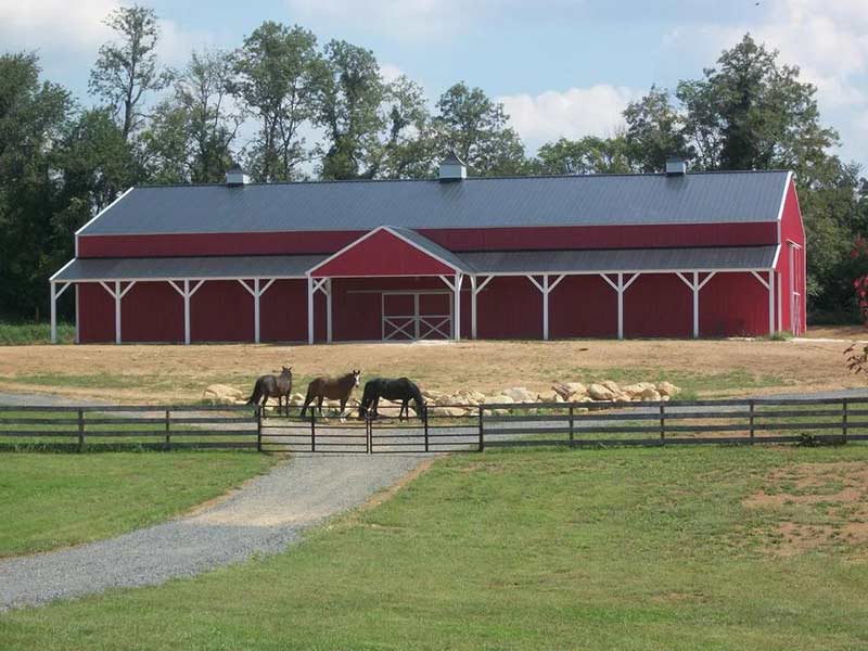 Long red horse barn with a center gable behind a fenced pasture with horses