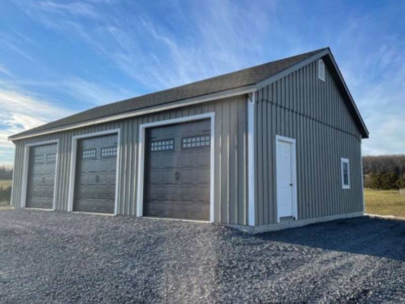 Gray detached garage with three carriage-style overhead doors on a gravel pad